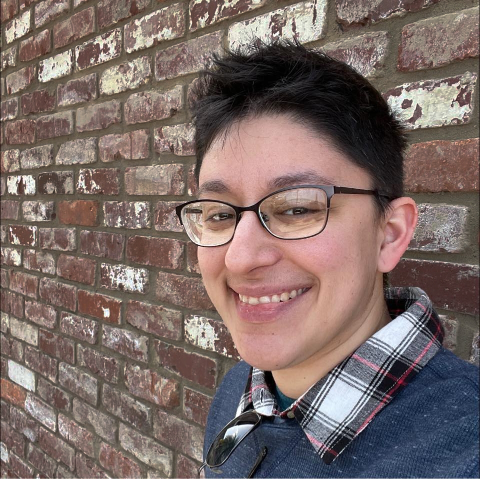 Danielle Zarcaro from the shoulders up, leaning against a brick wall with glasses, short dark hair, and a plaid button-up under a long sleeve shirt.